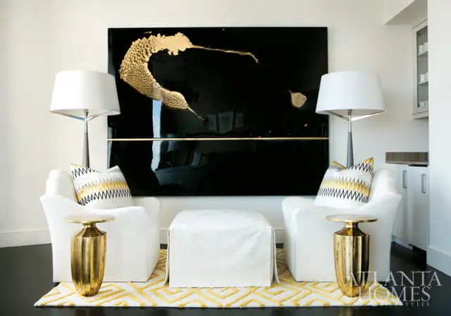 Two white armchairs flank a white ottoman, each with a gold accent table. A large black artwork with gold abstract splashes hangs on the wall above.