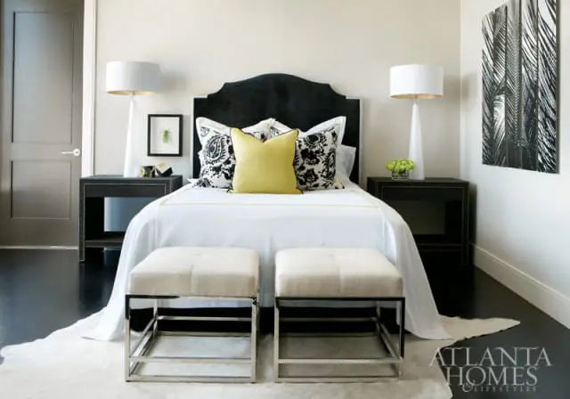 Bedroom with a black upholstered headboard, white bedding, yellow and black patterned pillows, and two cream upholstered benches with chrome frames.