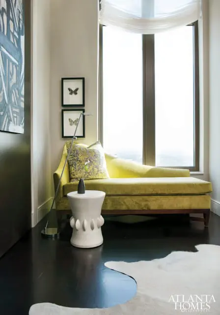 Lime green velvet chaise lounge with decorative pillows next to a white ceramic garden stool and framed butterfly art. Dark wood floors and a large window with sheer curtains.