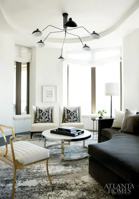 Living room with cream armchairs, black and white patterned pillows, a white coffee table with books, and a dark sectional sofa, all on a grey and white abstract rug.