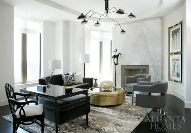 Modern living room with dark sofa, grey armchairs, brass coffee table, marble fireplace, and abstract rug. Atlanta Homes & Lifestyles logo in lower right corner.