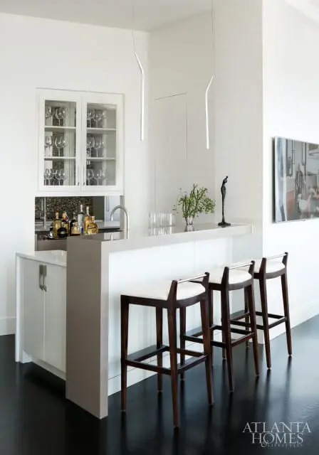 White bar area with glass-fronted cabinet displaying wine glasses, a quartz countertop with bottles, and three dark wood bar stools with white cushions.