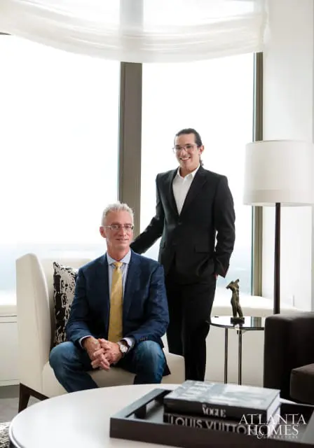 Two men in suits stand and sit in a modern living room with large windows. One man sits in a cream armchair wearing a blue plaid suit jacket, yellow tie, and jeans. The other man stands behind him, wearing a black suit and glasses.