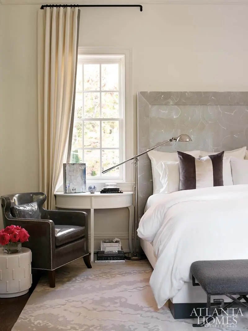 Bedroom with a large white bed, grey upholstered headboard, leather armchair, and a white side table next to a window.