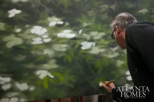 Artist painting a canvas with white flowers and green leaves, with the Atlanta Homes & Lifestyles logo in the lower right corner.