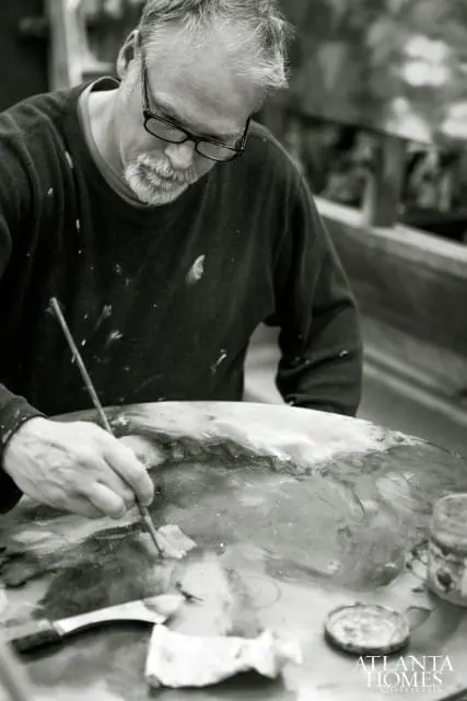 Man with glasses and beard painting at a palette, with paint splatters on his shirt.