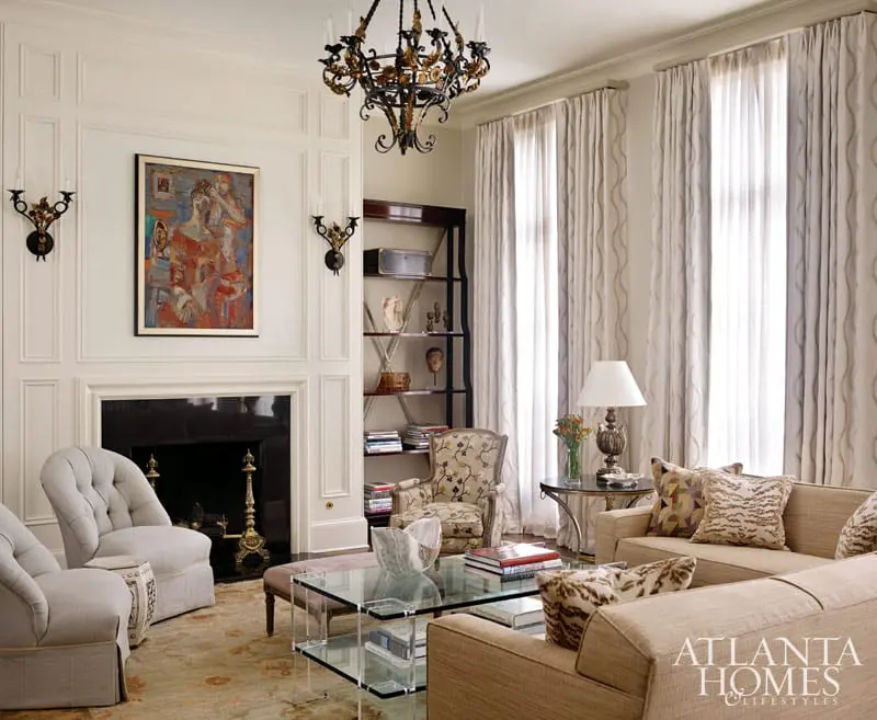 Living room with a fireplace, tufted armchairs, a glass coffee table, and a sofa. A large abstract painting hangs above the fireplace.