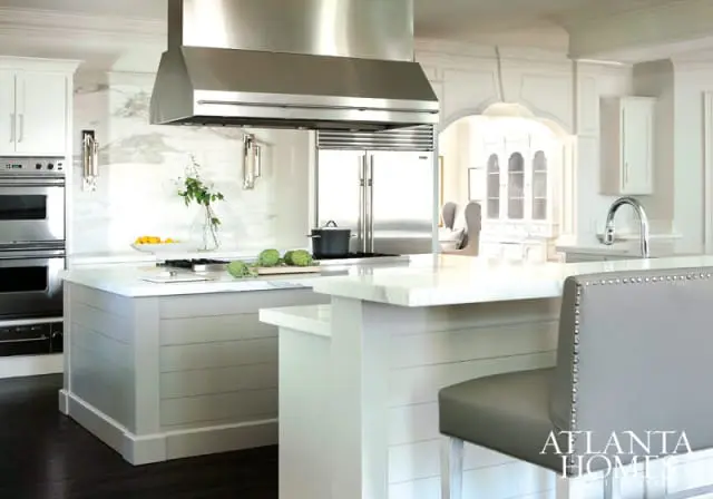 White kitchen with marble countertops, stainless steel range hood, shiplap island base, and a gray upholstered bar stool.