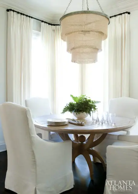 Round wooden dining table with white slipcovered chairs, a tiered burlap chandelier, and a large fern centerpiece.