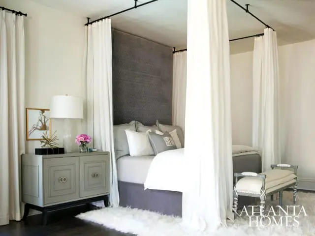 Bedroom with a four-poster bed draped in white curtains, a grey upholstered headboard, and a plush white rug. A grey cabinet with gold hardware sits beside the bed.