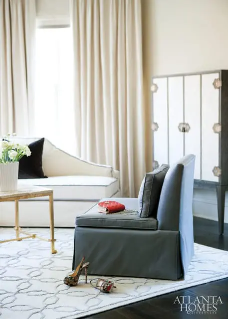 Gray upholstered chair with a red quilted clutch and jewelry on the seat, in front of a white chaise lounge and a tall cabinet with decorative hardware.