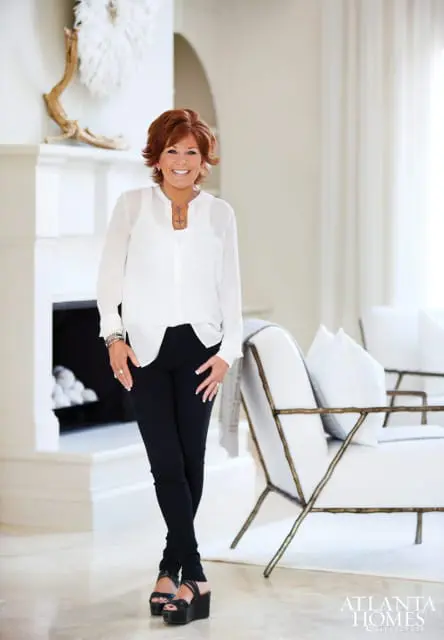 Woman with auburn hair smiling, wearing a white sheer blouse and black pants, standing in a well-lit living room with a fireplace and modern chairs.