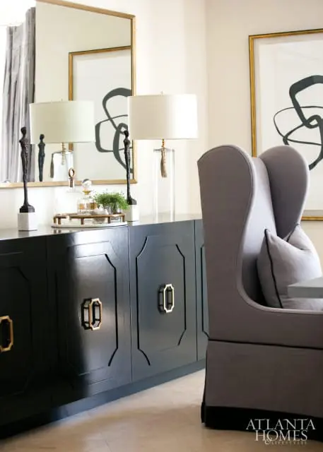 Dark blue sideboard with gold hardware, topped with two lamps, decorative bottles, and sculptures, next to a gray wingback chair.