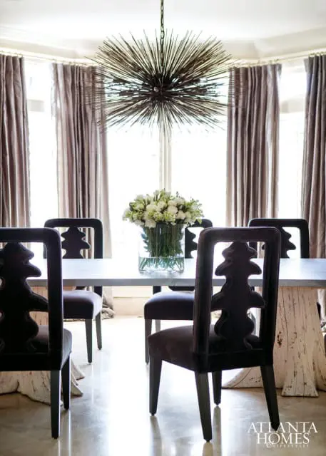 Dining room with a metal table, dark upholstered chairs with carved backs, and a sputnik chandelier. A vase of white flowers sits on the table.