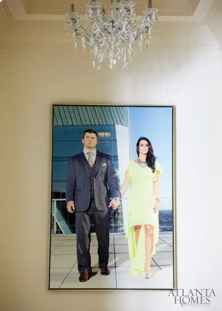A framed portrait of a man in a pinstripe suit and a woman in a lime green dress holding hands, standing on a balcony with a modern building in the background. A crystal chandelier hangs above.