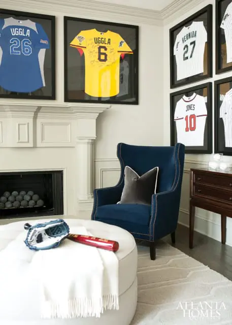 Living room with framed baseball jerseys, a blue wingback chair, white ottoman with baseball glove and bat, and a fireplace.