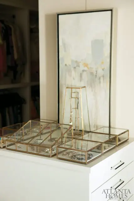 White dresser with glass and brass jewelry boxes filled with necklaces and bracelets. A tall glass jewelry stand and abstract art hang in the background.