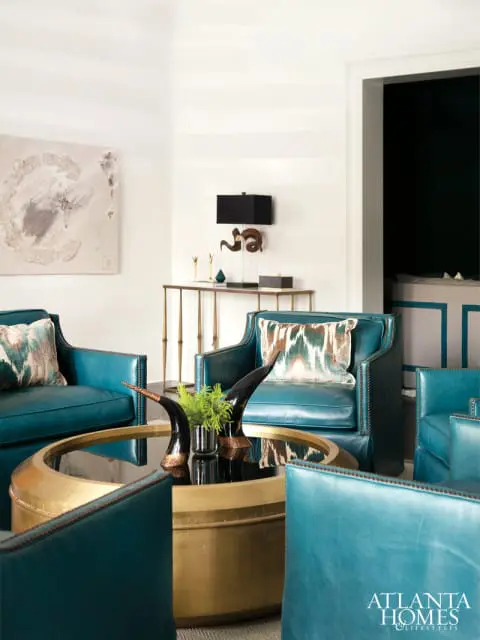 Living room with teal leather armchairs surrounding a brass and glass coffee table. Abstract art hangs on the wall.