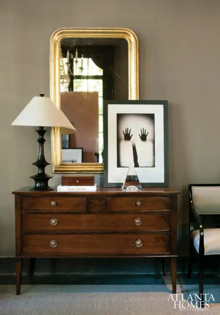 Dark wood dresser with brass hardware topped with a lamp, framed artwork of hands, a book, and a glass pyramid. A gold mirror hangs above.