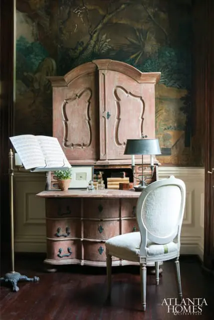Antique pink armoire with ornate doors and drawers, a desk with sheet music and a lamp, and a light upholstered chair in front of a mural-style wallpaper.