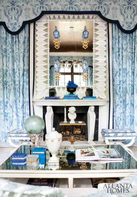 Blue and white patterned wallpaper and curtains frame a fireplace with a white mantelpiece, busts, and decorative vases. A mirrored coffee table holds books and decorative objects.