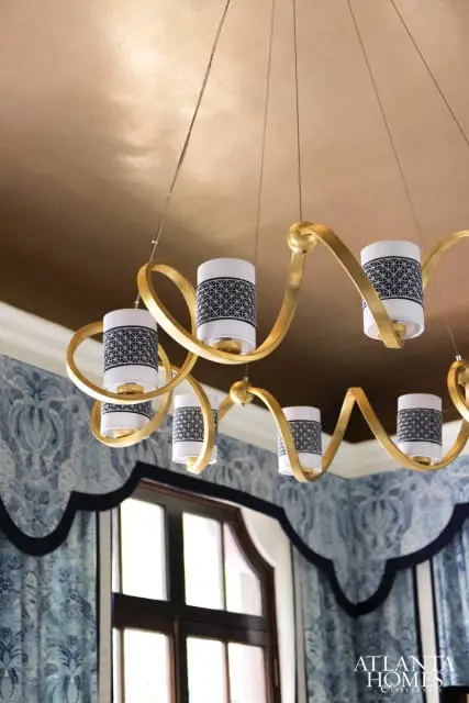 Gold chandelier with cylindrical white shades featuring a black geometric pattern hangs above a window with blue damask wallpaper.