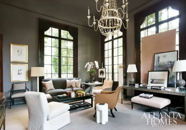 Living room with dark gray walls, a large ornate chandelier, a dark sofa with blush pillows, and a black coffee table. Large windows with dark frames overlook greenery.