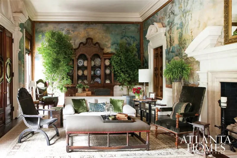Living room with a large wooden hutch filled with plates, a white sofa with green pillows, and a long upholstered ottoman. The walls feature a scenic mural and a fireplace.