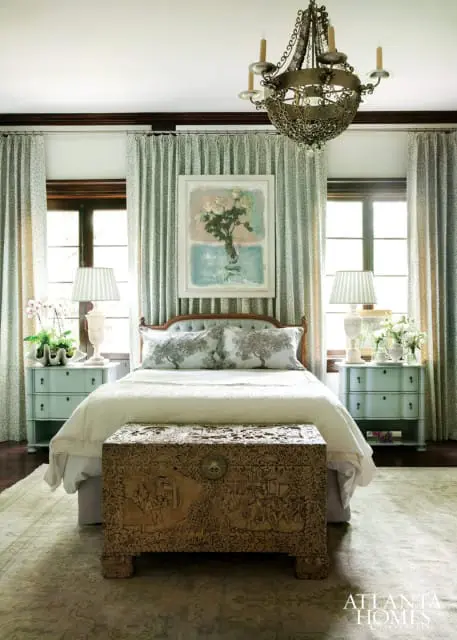 Bedroom with a light blue tufted headboard, white bedding, and decorative pillows featuring tree motifs. A carved wooden chest sits at the foot of the bed.