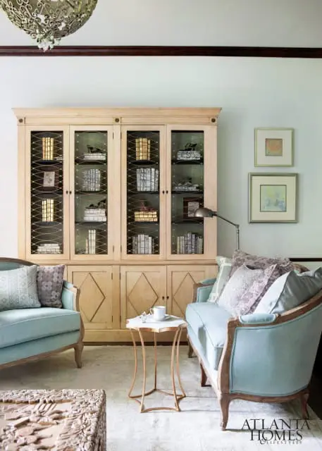 Light wood built-in cabinet with glass doors displaying books and decorative items, flanked by two light blue upholstered armchairs and a small marble-topped side table.