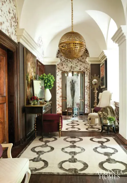 Hallway with vaulted ceiling, floral wallpaper, dark wood doors, console table with artwork and flowers, a burgundy ottoman, and a large chain-link patterned rug.