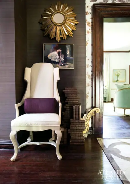 White upholstered armchair with a purple bolster pillow, next to a stack of antique books. A sunburst mirror and painting hang on the dark textured wall.