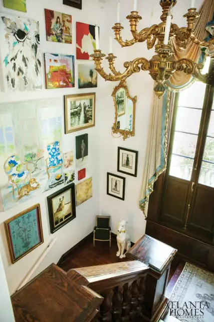 Hallway with a collection of diverse artwork on white walls, a gilded chandelier, a mirrored sconce, and a large wooden staircase with ornate railings.