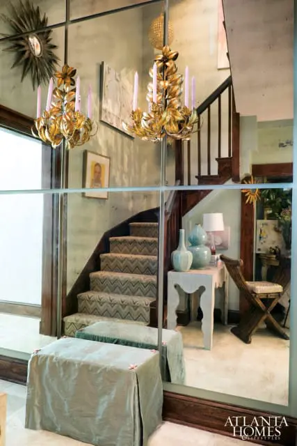 Hallway with mirrored walls, a chevron-patterned carpeted staircase, ornate gold chandeliers with pink candles, and a sunburst mirror.