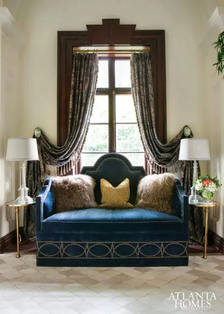 Deep blue velvet sofa with ornate gold trim and plush pillows in front of a dark wood-trimmed window with patterned drapes.