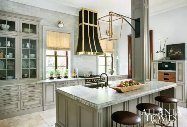 Gray kitchen with marble island, black range hood with gold accents, glass-front cabinetry, and pendant light.
