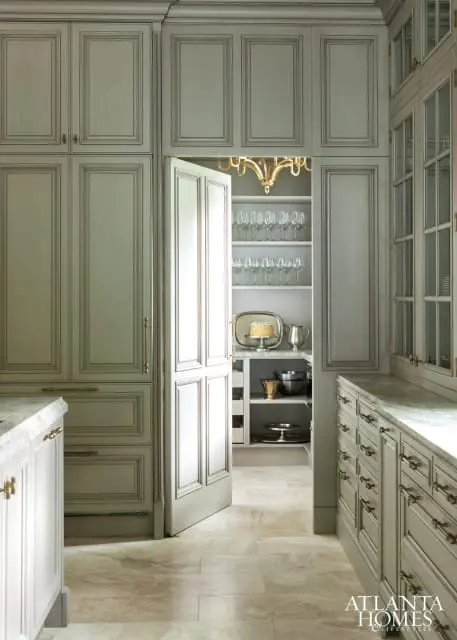 Light gray kitchen with paneled cabinetry, marble countertops, and a walk-in pantry with shelves of glassware and a cake on a silver tray.