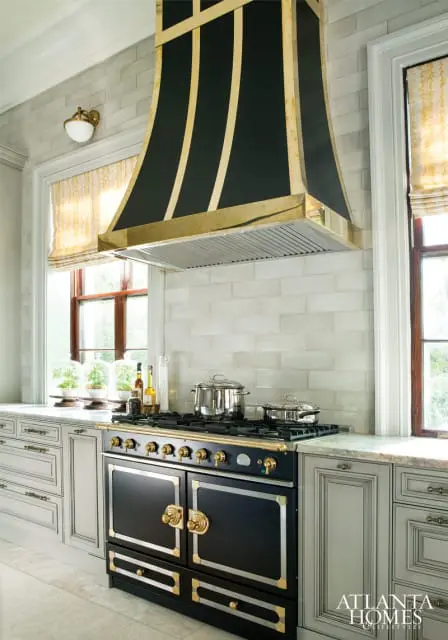 Black and brass range hood over a black range with gold accents, set against a light gray subway tile backsplash. Light gray cabinetry flanks the range.