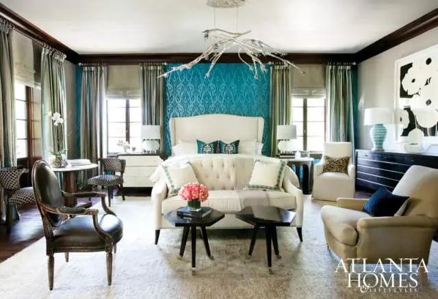 Bedroom with a cream tufted wingback bed, teal patterned wallpaper, cream sofa, and matching armchairs. A branch-like chandelier hangs overhead.