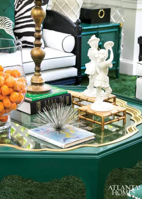 Green lacquered coffee table with mirrored top displaying books, a glass vase of tangerines, a brass lamp, and white figurines on bamboo stands.