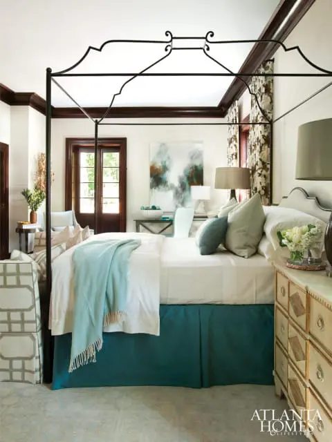 Bedroom with a black canopy bed, white bedding, teal bed skirt, and a patterned armchair. A dresser with ornate detailing is to the right.