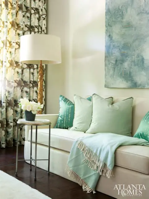 Cream sofa with light green and teal patterned pillows, a light blue throw blanket, and a gold floor lamp next to floral curtains.