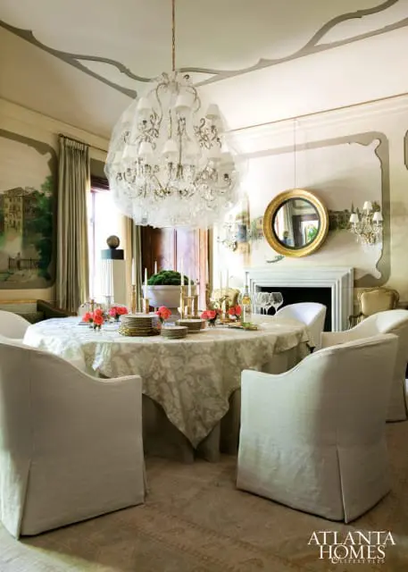 Dining room with a round table covered in a damask tablecloth, set with gold-rimmed plates and coral flowers. Upholstered chairs surround the table, and a large crystal chandelier hangs from the ceiling.