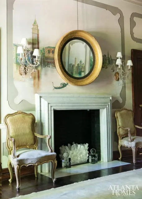 Ornate gold-framed mirror above a marble fireplace with a Venetian mural on the wall. Two antique chairs flank the fireplace.