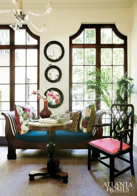 Sunlit sitting area with a blue upholstered chaise lounge, floral and leopard print pillows, a marble-topped pedestal table, and a dark wood chair with a coral seat.