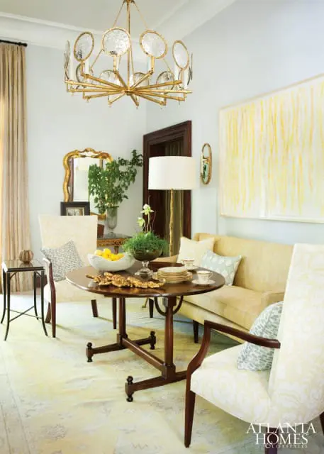Light yellow living room with a round wooden dining table, a pale yellow sofa, upholstered chairs with a floral pattern, and a large abstract painting with yellow and white brushstrokes.