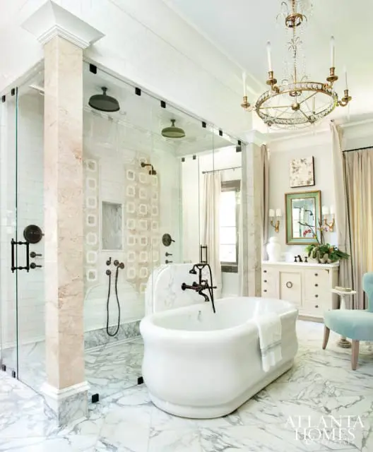 Luxurious bathroom with a freestanding white tub, marble floors, and a glass-enclosed shower with multiple showerheads. A vintage-style chandelier hangs overhead.