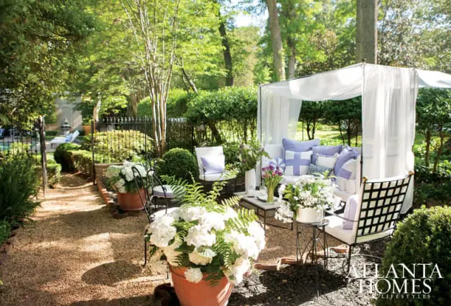 Outdoor seating area with a white canopy bed, lavender and white pillows, and lush greenery. A gravel path leads to the seating area, with potted white hydrangeas in the foreground.
