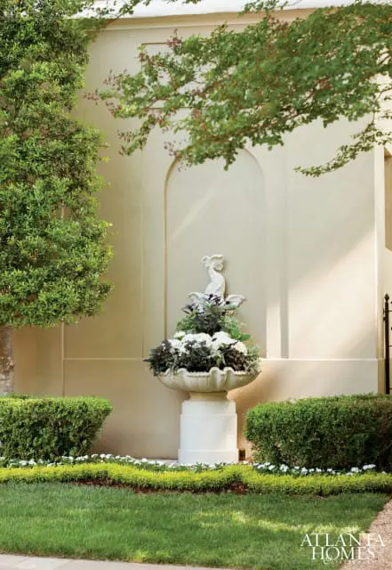 Stone urn filled with white and dark foliage plants, topped with a white sculpted bird, set against a stucco wall with an arched niche. Trimmed hedges and a manicured lawn surround the urn.