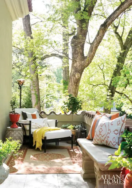 Covered porch with white outdoor sofa and chaise, stone bench, patterned rug, and lush green trees in the background.